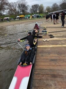 Rowing team prepares for practice on a cloudy day, with team members and equipment on the water's edge.