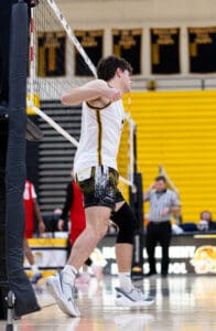 Male volleyball player celebrates after a point during a high school match, with teammates and referees in the background.