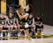High school volleyball player serving the ball, teammates watching from the bench in a gym with yellow and black colors.