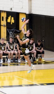 High school volleyball player serving the ball, teammates watching from the bench in a gym with yellow and black colors.