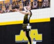 High school volleyball player jumping to serve during a match in a gym with team banners in the background.