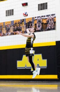 High school volleyball player jumping to serve during a match in a gym with team banners in the background.