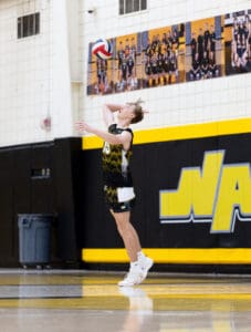 A volleyball player prepares to serve during a match in a gym with team photos in the background.
