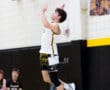A young male volleyball player jumps to serve a ball during a game, with teammates in the background.