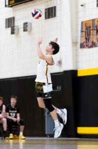 A young male volleyball player jumps to serve a ball during a game, with teammates in the background.