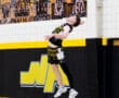 High school volleyball player performing a powerful jump serve in a gym decorated with team photos.
