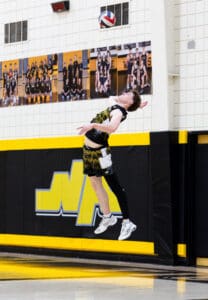 High school volleyball player performing a powerful jump serve in a gym decorated with team photos.