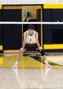 High school volleyball player in action stance, focused on the game, inside a gym with a yellow and black theme.