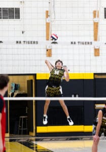 Boy in a volleyball uniform jumping to spike a ball over the net during a game in a gymnasium.