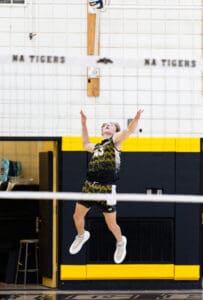 High school volleyball player jumps to serve the ball in an indoor gym setting, showcasing athletic skill and focus.