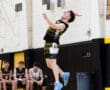 A player leaps to serve during a volleyball match, teammates cheering from the bench in the background.
