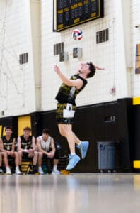 A player leaps to serve during a volleyball match, teammates cheering from the bench in the background.
