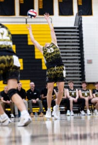 A volleyball player leaps to serve during a game, with teammates on the bench in the background and a vibrant gym setting.