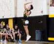 A young volleyball player jumps to serve during a game, with teammates watching from the bench.