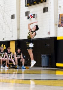 A young volleyball player jumps to serve during a game, with teammates watching from the bench.