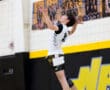 A volleyball player jumping to serve, focused and airborne in a gym with team photos in the background.