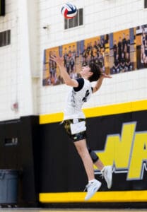 A volleyball player jumping to serve, focused and airborne in a gym with team photos in the background.