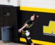 Young male volleyball player executing a jump serve in a gym with a yellow and black color scheme.