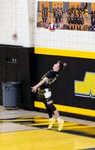 Young male volleyball player executing a jump serve in a gym with a yellow and black color scheme.