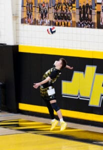 Young volleyball player jumping to serve a ball in a gymnasium with black and yellow decor, showcasing athleticism.