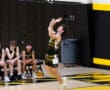 High school volleyball player serves the ball while teammates watch from the sidelines in a gym with yellow and black colors.