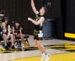 High school volleyball player jumping to serve, with teammates in background on a gym court featuring yellow and black colors.