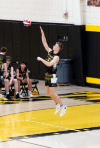 High school volleyball player jumping to serve, with teammates in background on a gym court featuring yellow and black colors.