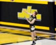 A male volleyball player serves the ball during a game on a gym court with yellow and black decorations.