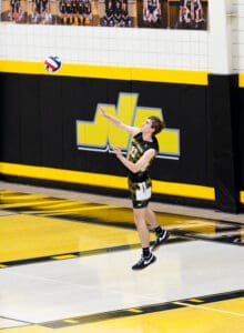 A male volleyball player serves the ball during a game on a gym court with yellow and black decorations.