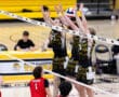 Players jump to block a volleyball in a competitive match, with fans in the background at a school gym.