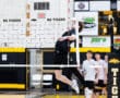 High school volleyball player jumps to spike the ball over the net during a game, teammates in the background.