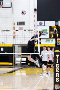 High school volleyball player jumps to spike the ball over the net during a game, teammates in the background.