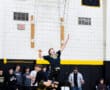 Male volleyball player jumping to serve during a practice session in a school gymnasium, with teammates observing.
