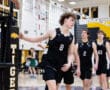 High school volleyball players in black jerseys play during a match, with a referee officiating at the net.