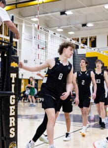 High school volleyball players in black jerseys play during a match, with a referee officiating at the net.