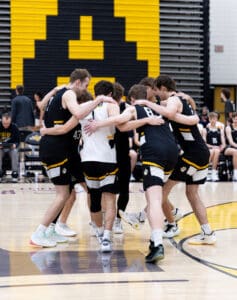 Basketball team huddled together, showing teamwork and strategy before their game on a court with black and yellow colors.