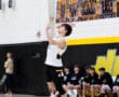 High school athlete serving volleyball during a match, with teammates observing on the sidelines.