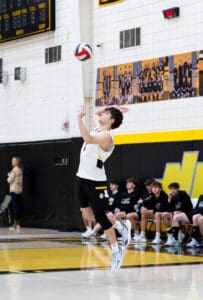 High school athlete serving volleyball during a match, with teammates observing on the sidelines.