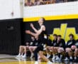 A volleyball player prepares to serve, with teammates sitting on the bench in the background at a school gymnasium.