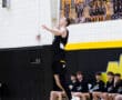 Player in a black volleyball jersey jumps to serve during a game, with teammates watching from the bench.