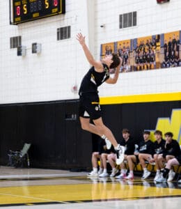 High school volleyball player jumping to serve during a match, with teammates cheering on the sidelines.