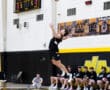 High school volleyball player jumps to serve during a match, with teammates and score visible in the background.