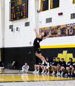 High school volleyball player jumps to serve during a match, with teammates and score visible in the background.