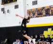 High school volleyball player serves the ball during a match, with teammates watching in the background.