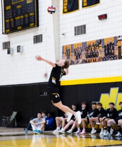 High school volleyball player serves the ball during a match, with teammates watching in the background.