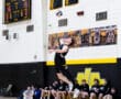 High school volleyball player jumps to serve, with teammates cheering on the sideline and score displayed above.