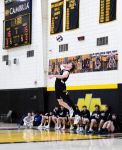 High school volleyball player jumps to serve, with teammates cheering on the sideline and score displayed above.