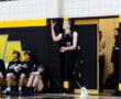 Volleyball player serves the ball during a match, with teammates seated on the bench in the background.