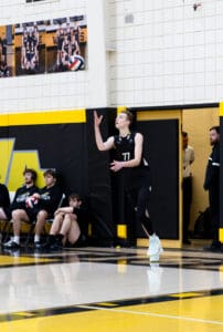 Volleyball player serves the ball during a match, with teammates seated on the bench in the background.