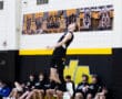 Athlete performing a volleyball serve in gym, teammates watching from the sidelines. Energetic team sport moment captured.
